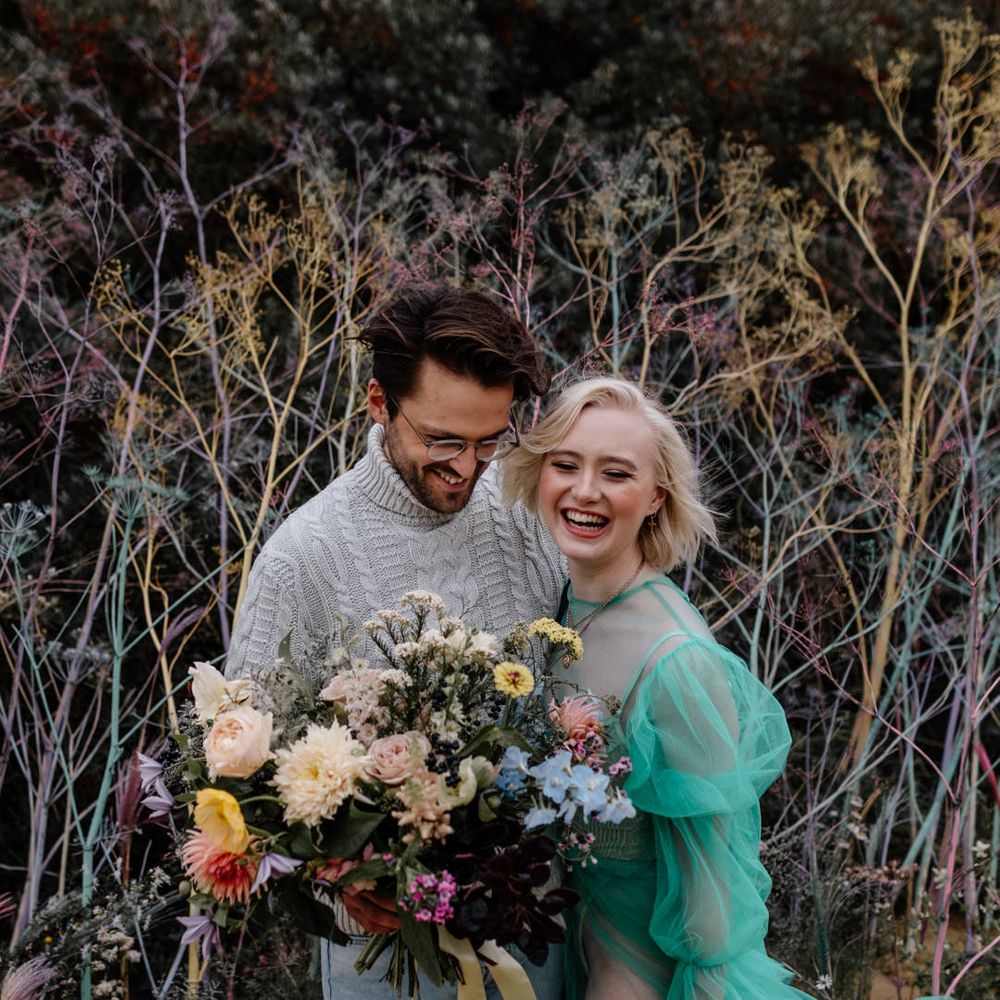Bride and groom laughing at their beach elopement standing infant of a sand dune with colourful flower stems 