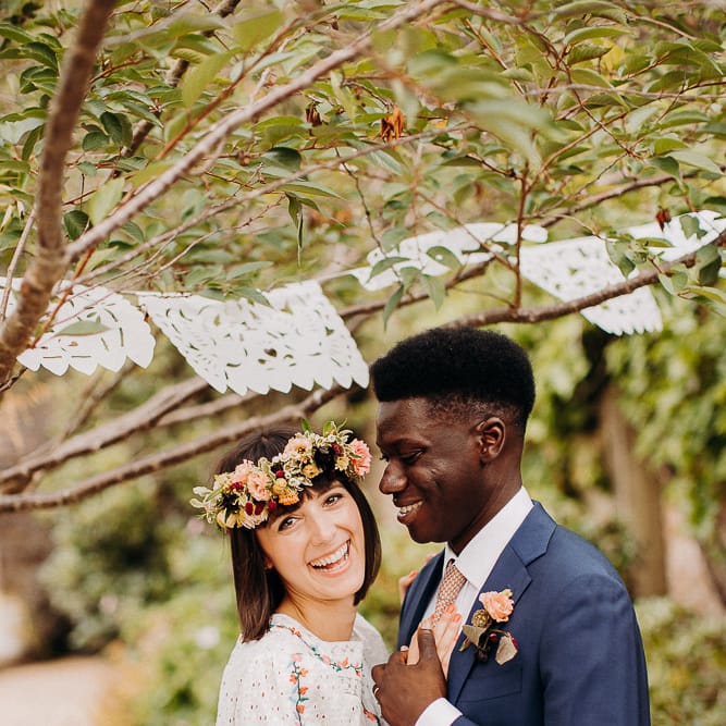 Groom in a navy blue suit embracing his bride in a Broderie Anglaise wedding dress under a canopy of laser cut bunting