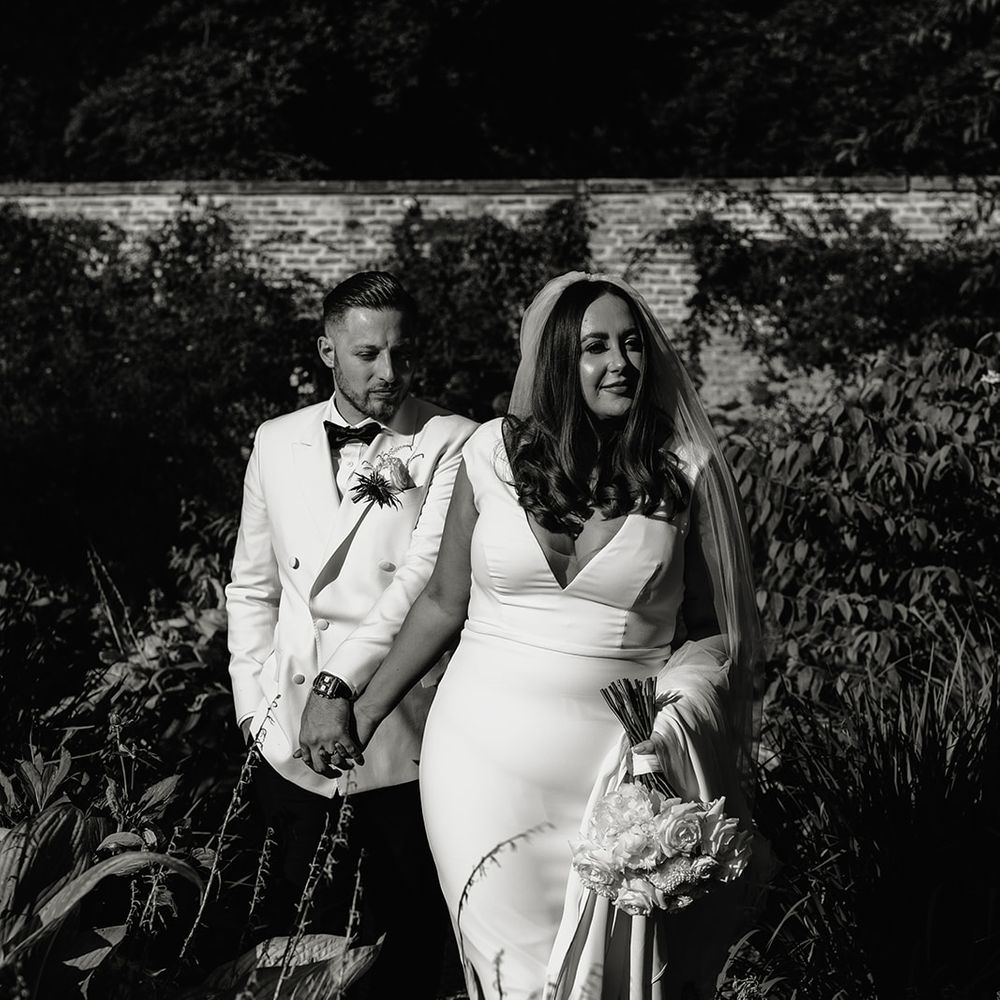 Bride in V neck Made With Love wedding dress and veil holding white rose bouquet walks outside with groom in white double breasted tuxedo jacket