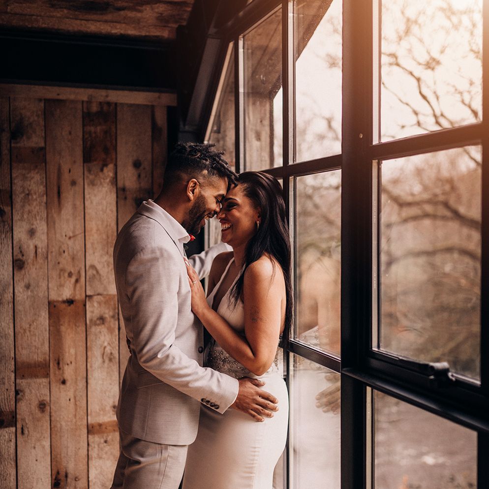 Bride & groom stand beside window as they look lovingly at one another 