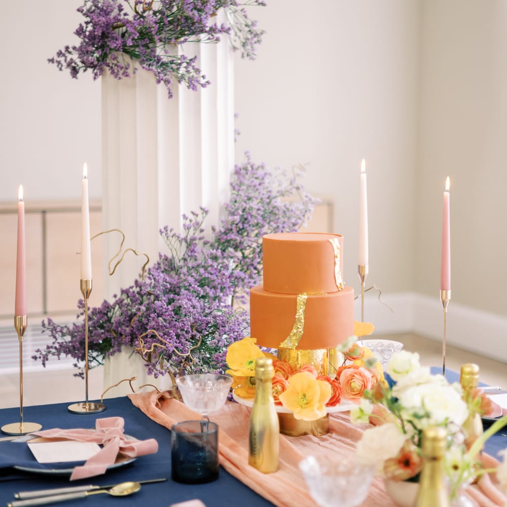 Orange frosted wedding cake complete with gold leaf and floral decor next to lavender display winding up pillar
