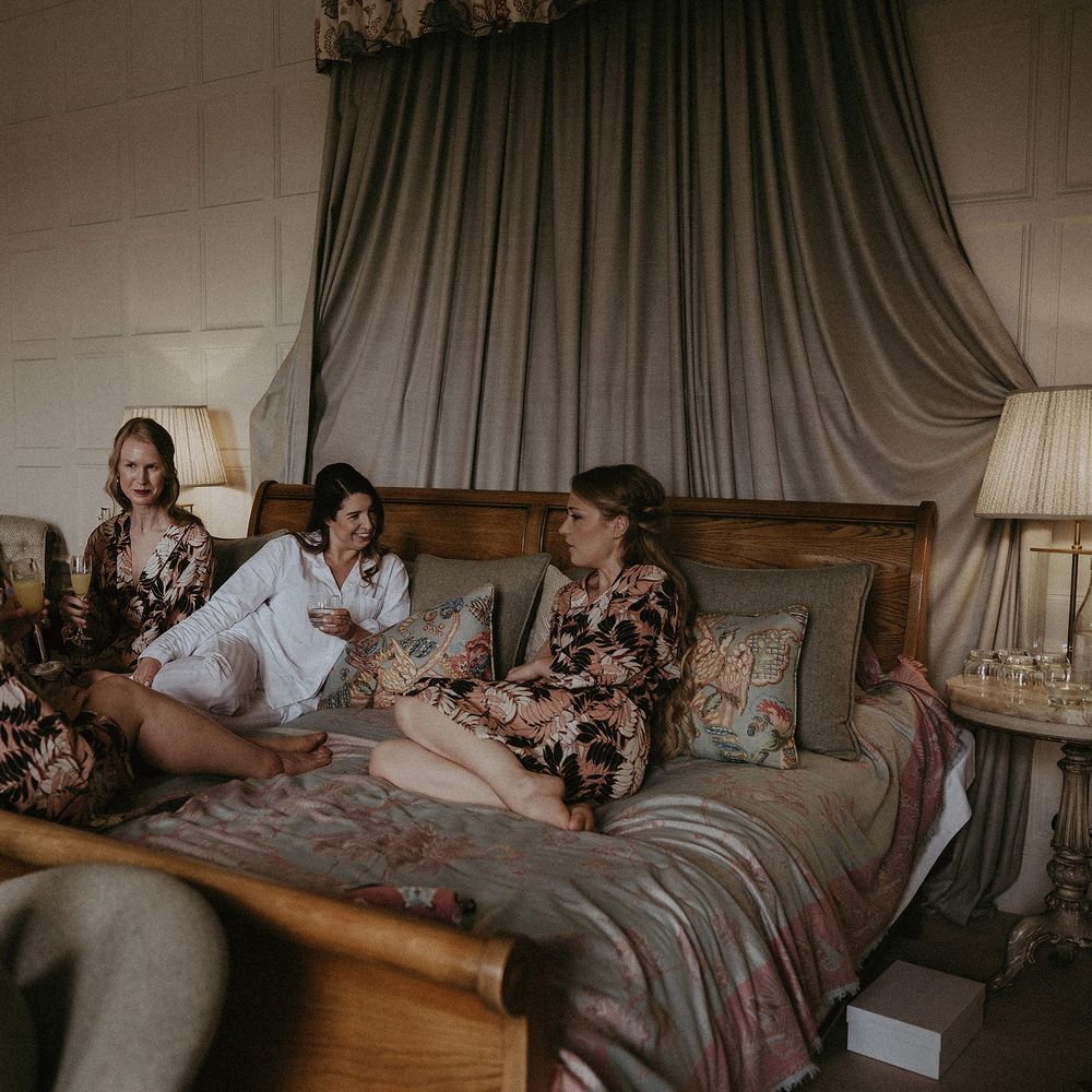Bride sits with her bridesmaids on the morning of her wedding as bridesmaids wear floral dressing gowns and drink champagne