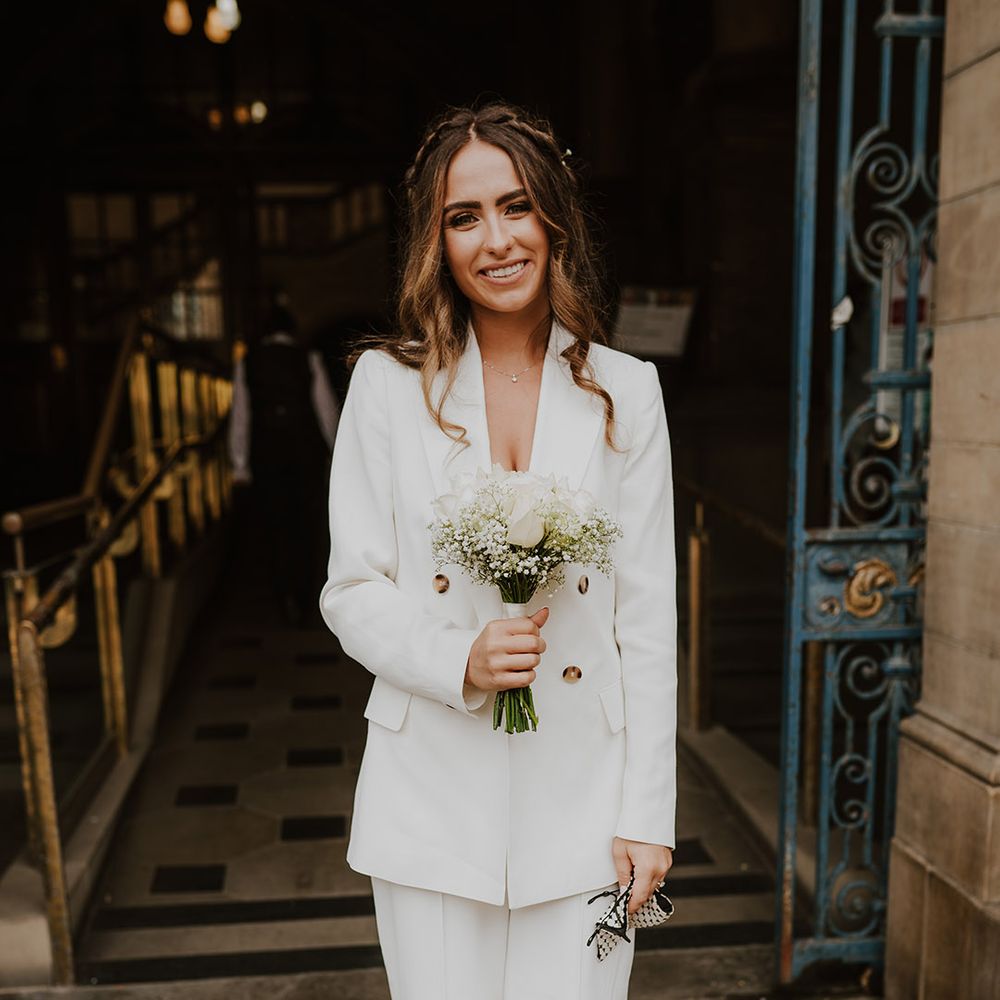 Bride in a white trouser suit and blazer holding a white rose and gypsophila bouquet 