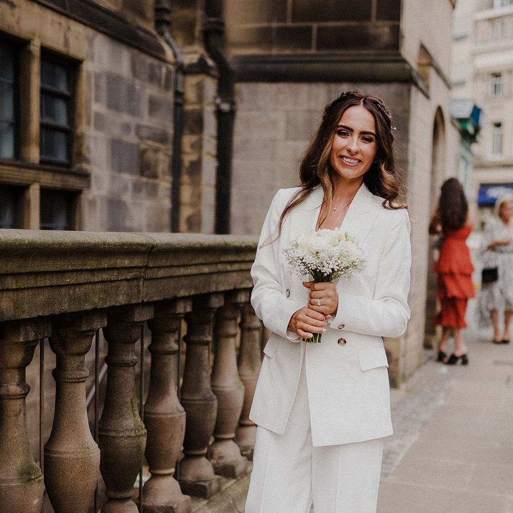 Stylish bride in a three piece white suit with jacket and wide leg trousers holding a white rose bouquet 