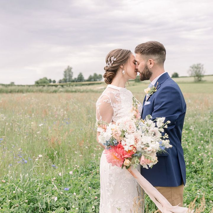 Groom in a navy blazer kissing his bride in a lace high street wedding dress in a field as she holds her wildflower wedding bouquet 