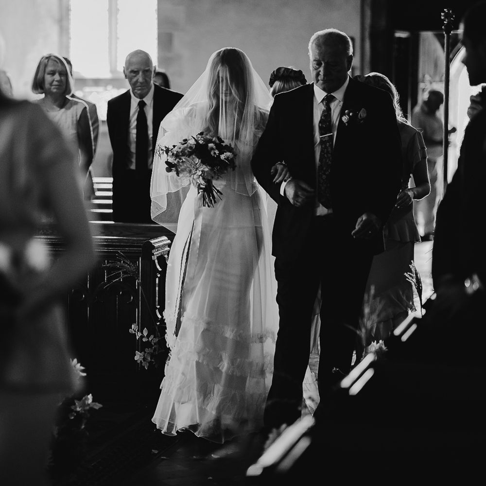 Bride in Halfpenny London Mayfair dress and veil holding mixed bridal bouquet walks down the aisle arm in arm with father in suit during church ceremony in Cornwall