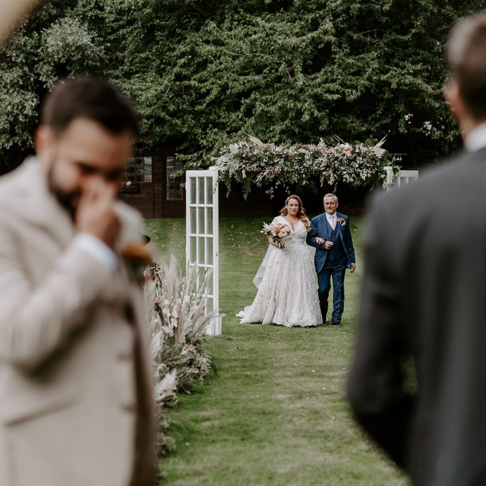 Bride in homemade wedding dress holding rose bridal bouquet walks up the aisle holding arm of man in blue three piece suit during DIY garden wedding ceremony in Bedfordshire
