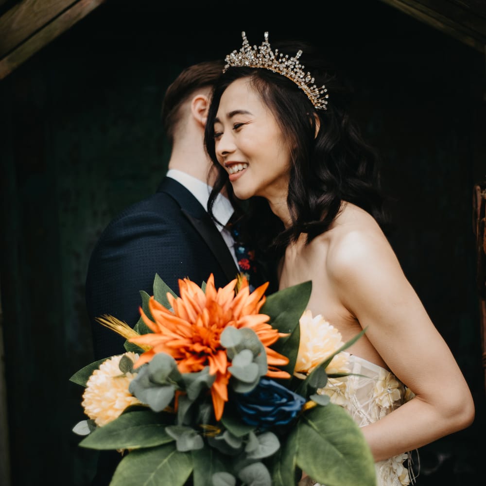 Smiling bride in headpiece and strapless wedding dress holds tropical wedding bouquet as groom in dark suit hugs her