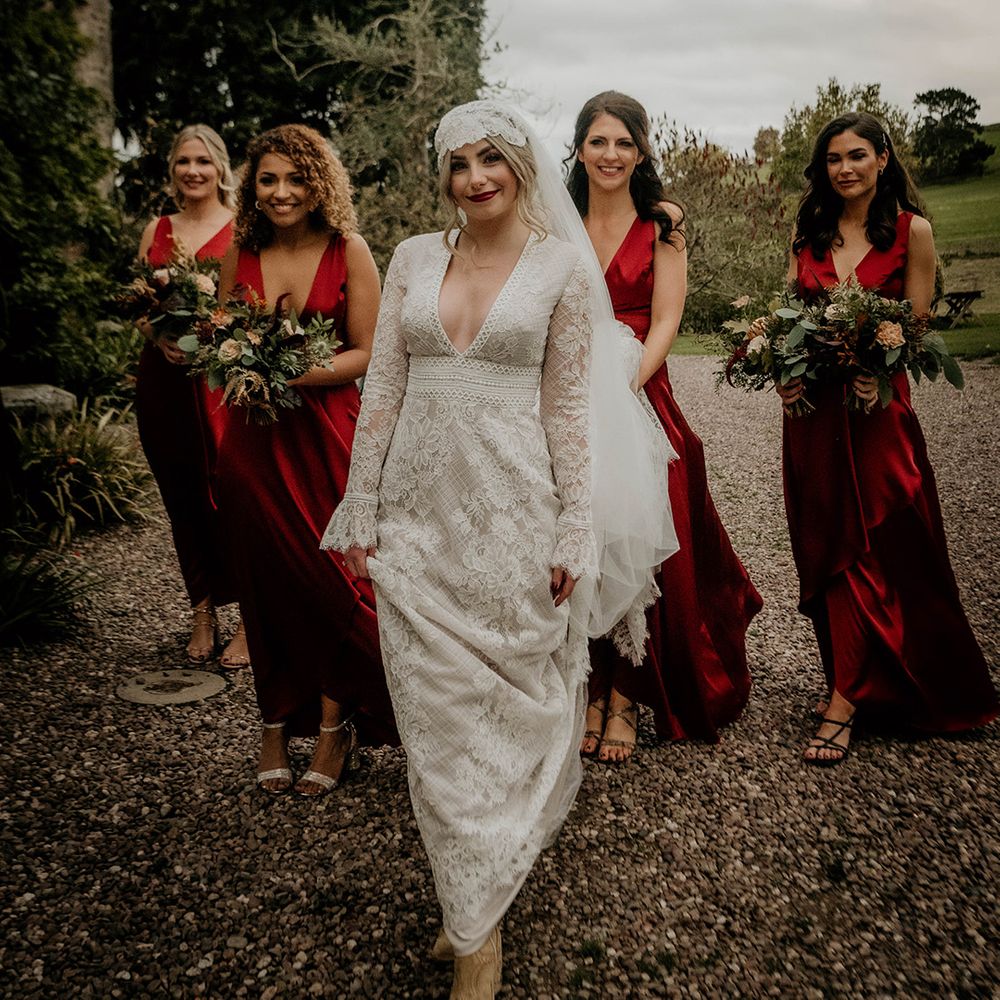 Boho bride in a lace wedding dress with plunging neckline and long lace bell sleeves standing with her bridesmaids in satin red dresses 