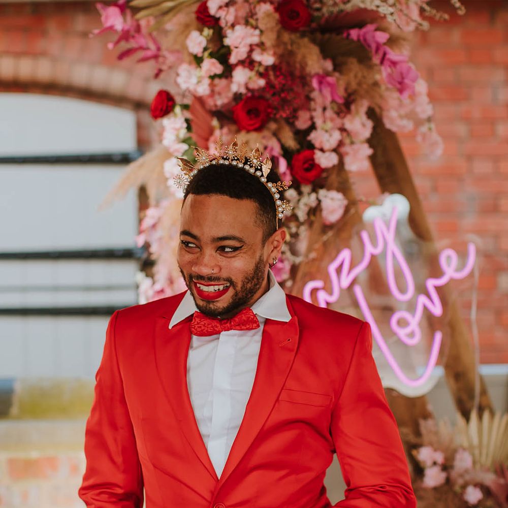 Groom in a red suit and bow tie wearing black winged eyeliner and a gold crown accessory 