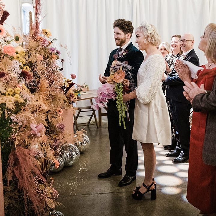 Wedding guests stand and clap as the bride and groom stand at the altar with the bride in a short wedding dress and from in velvet suit jacket 