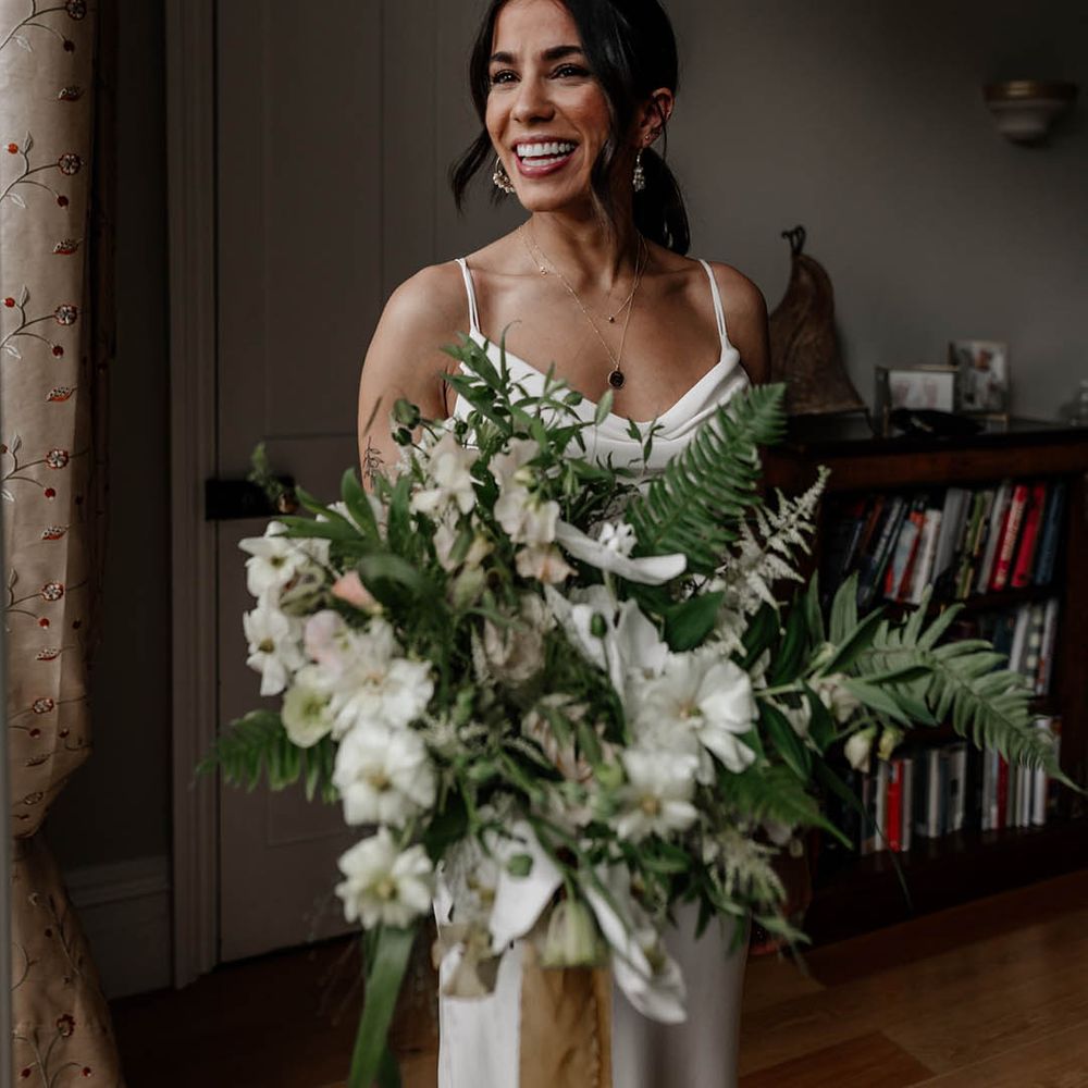Indian bride with natural makeup up and wavy ponytail wearing stacking necklaces and holding a white and green wedding bouquet 