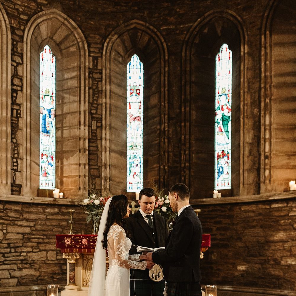 Bride wears floor length veil and holds the hands of her groom in front of stained glass windows during church ceremony 