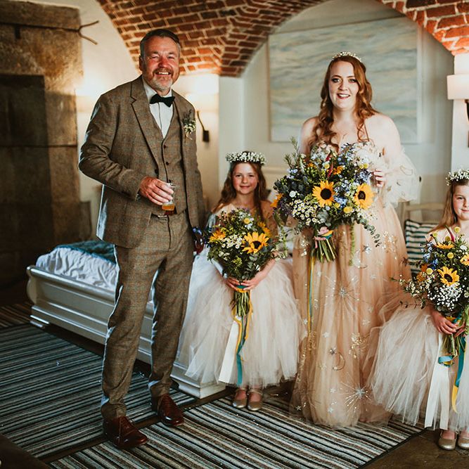 Bride wearing pink starry wedding dress stands beside flower girls who wear light pink dresses with ballerina skirts and floral crowns holding sunflower bouquets