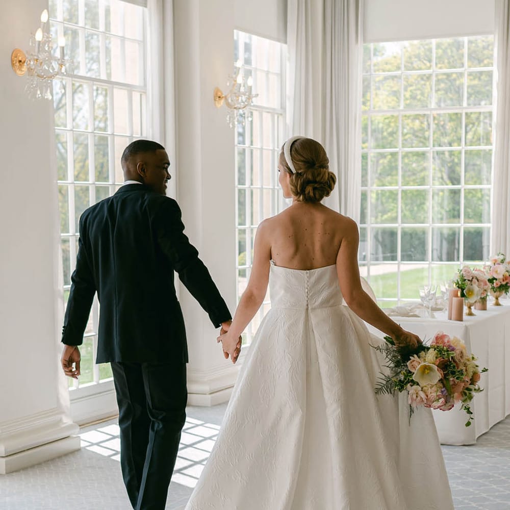 Bride wears a beaded headband with her updo hairstyle with a strapless brocade wedding dress with groom in black tie walk into their wedding breakfast