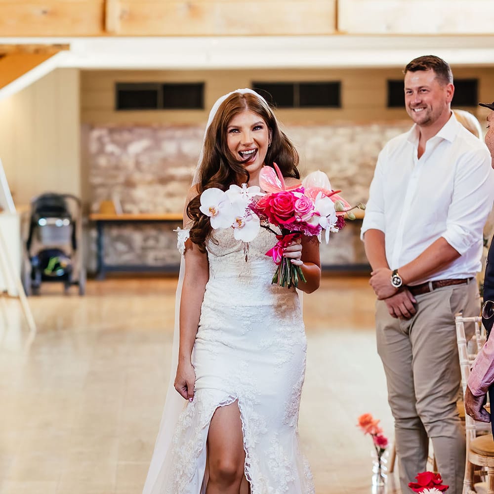 Bride walks down the aisle wearing floor length veil and off the shoulder lace wedding dress whilst holding bright pink floral bouquet 