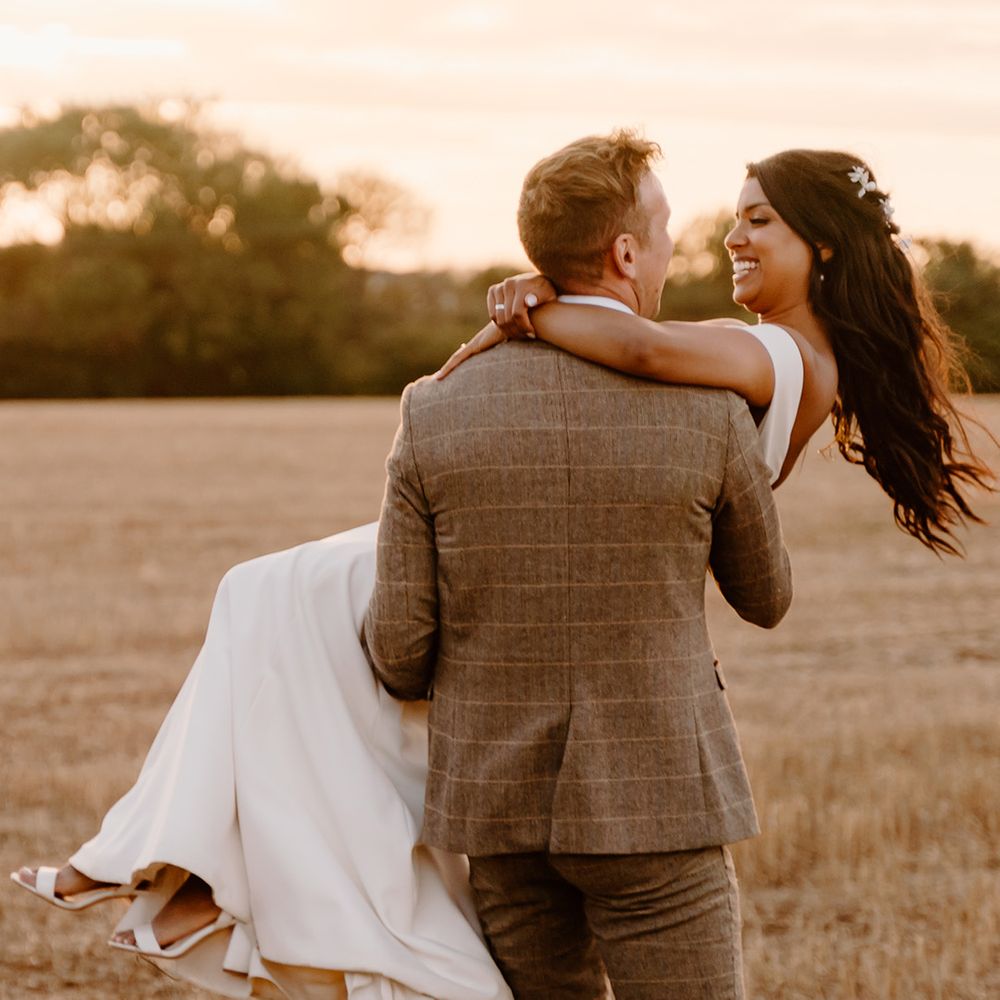 Groom carries the bride as they stand in a field together 