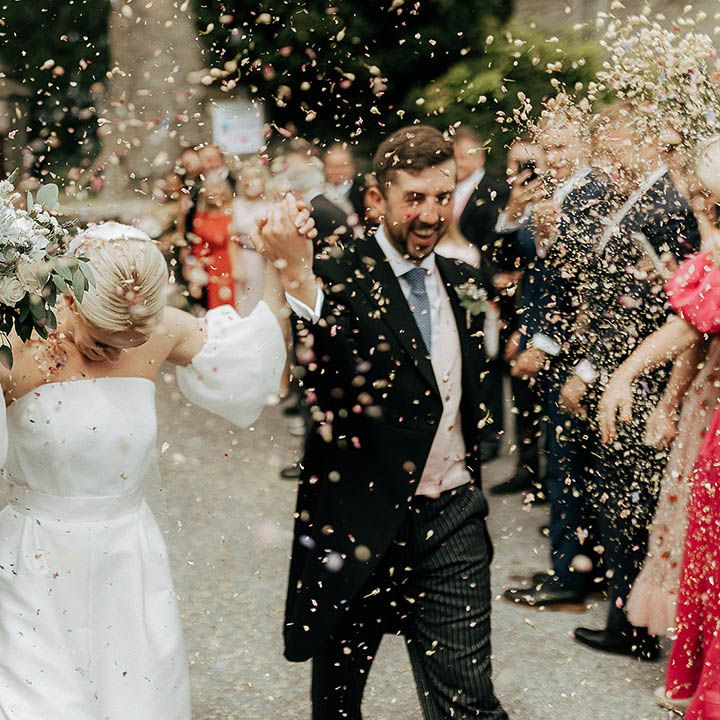 Bride & groom in morning suit and pale blue tie walk through colourful confetti exit 