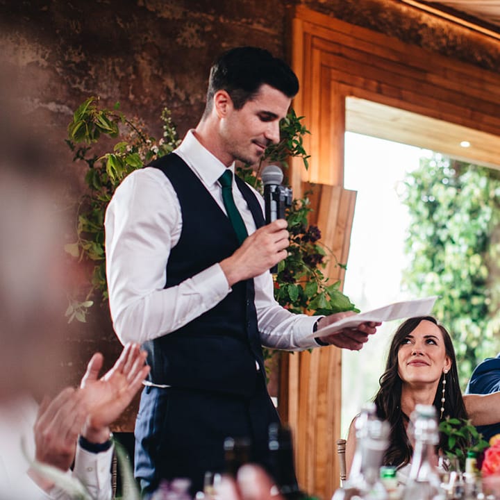 Groom stands up with a piece of paper and microphone to read out his wedding speech with the bride raising her glass in a toast 