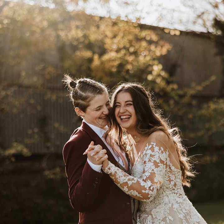 Bride in long sleeve lace illusion sleeve wedding dress with overlay dancing and laughing with bride in maroon bridal suit with grey waistcoat, dusky pink tie and autumnal dried flower boutonniere 