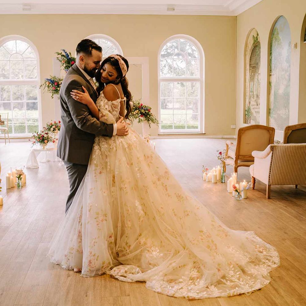 Bride in v-neck long sleeve shoulder cut out 3d appliqué wedding dress with flecks of gold wearing satin bridal headband dancing with groom in black blazer with pink garden rose and foliage boutonniere, cornflower blue waistcoat and black suit trousers 