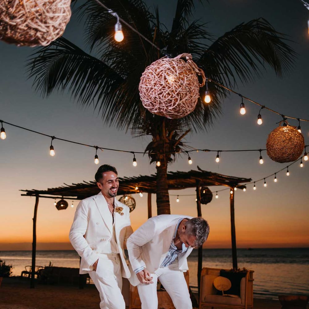 Groom in mens white wedding suit with dusky orange garden rose and dried flower boutonniere, white shirt and silver jewellery laughing with groom in white blazer, light blue and white striped shirt, dusky orange garden rose and dried flower boutonniere and gold jewellery 