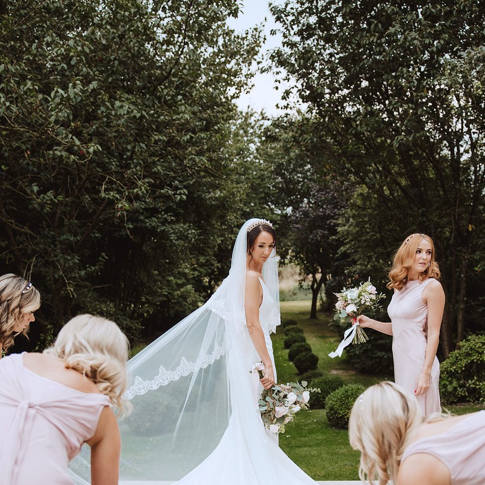Bride wearing cathedral length embroidered edge veil in elegant satin slip wedding dress surrounded by bridesmaids in pink dresses 