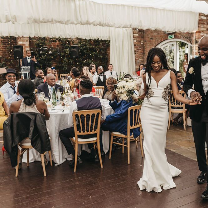 Bride in a fitted wedding dress with hip detail walking onto the dance floor with her groom in a black velvet tuxedo 