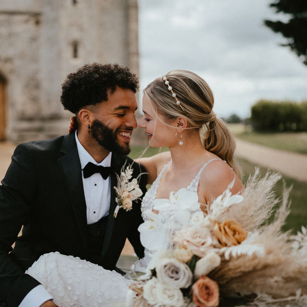 Groom in a three piece black suit and bow tie smiling at his bride with a sleek back ponytail and peal headband 