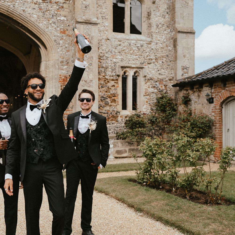 Groomsmen in tuxedos with the groom in a jacquard horseshoe waistcoat holding a bottle of champagne in the air 
