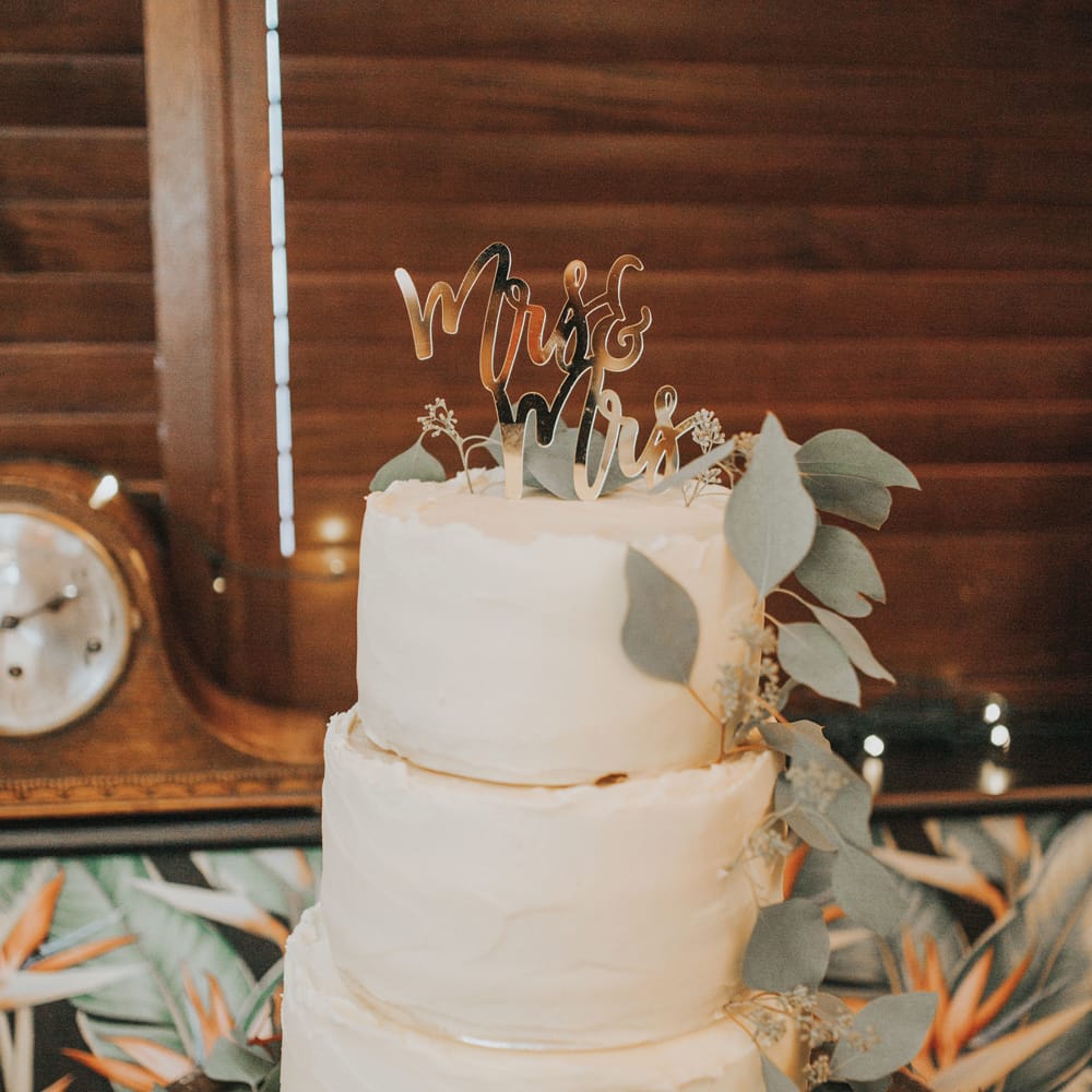 A three tiered white buttercream wedding cake has a topper reading mrs & mrs and is decorated with foliage at Crab & Lobster fairytale wedding.