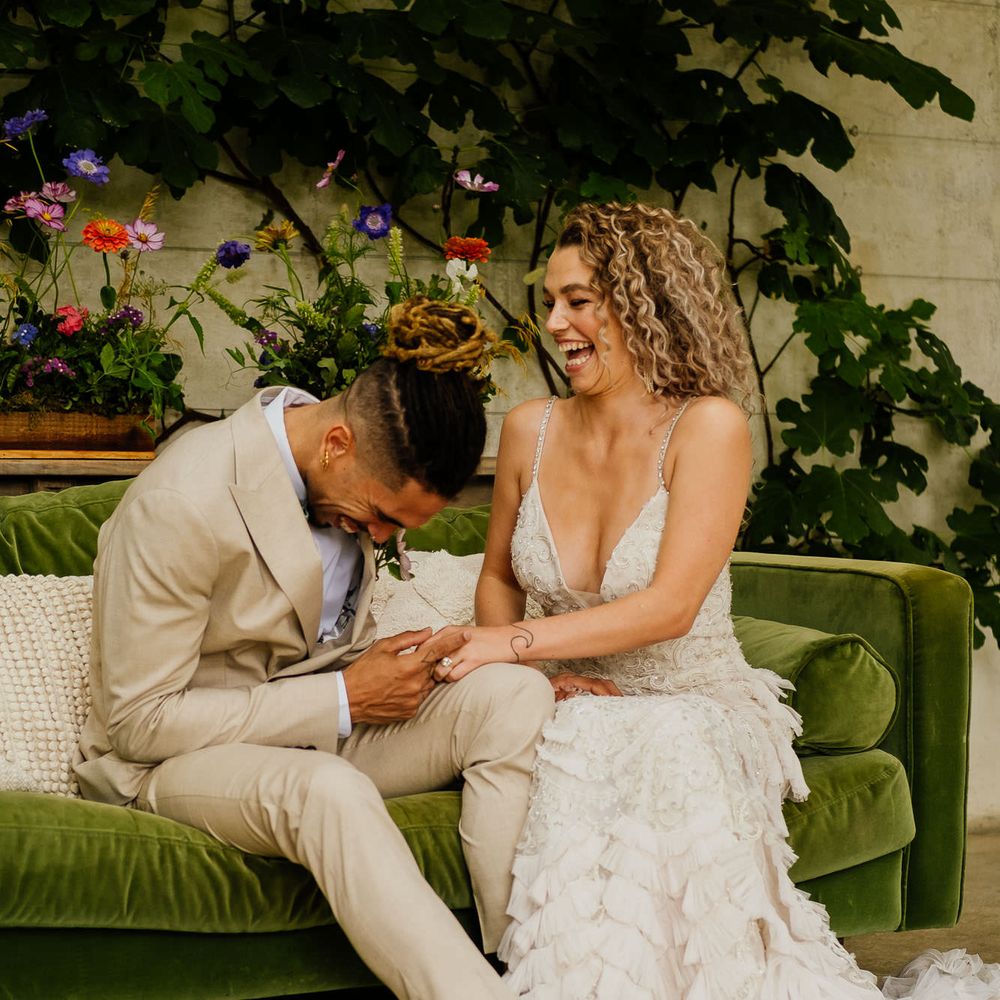 Groom in a beige suit and bride in an embellished wedding dress with straps and ruffles laughing on a green velvet sofa 