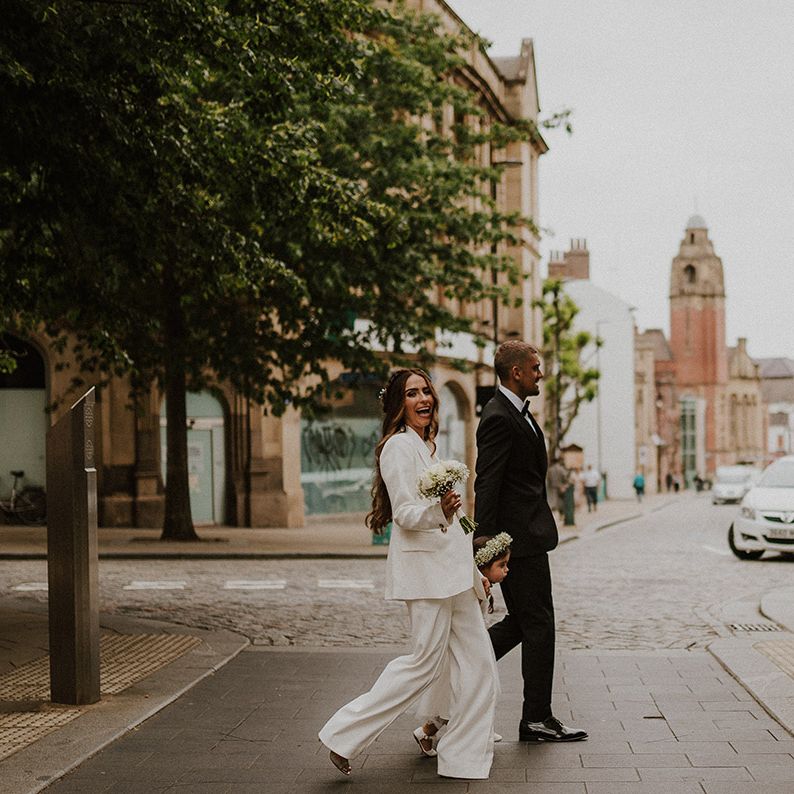 Bride in a wide leg trousers and blazer suit walking through Sheffield with her flower girl daughter and groom in a tuxedo 