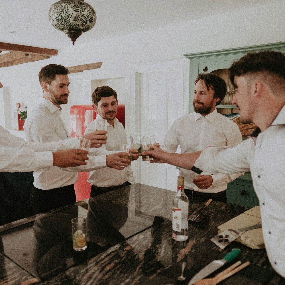 Grooms in white shits all clink glasses before Isle of Wight wedding with macrame wedding decor