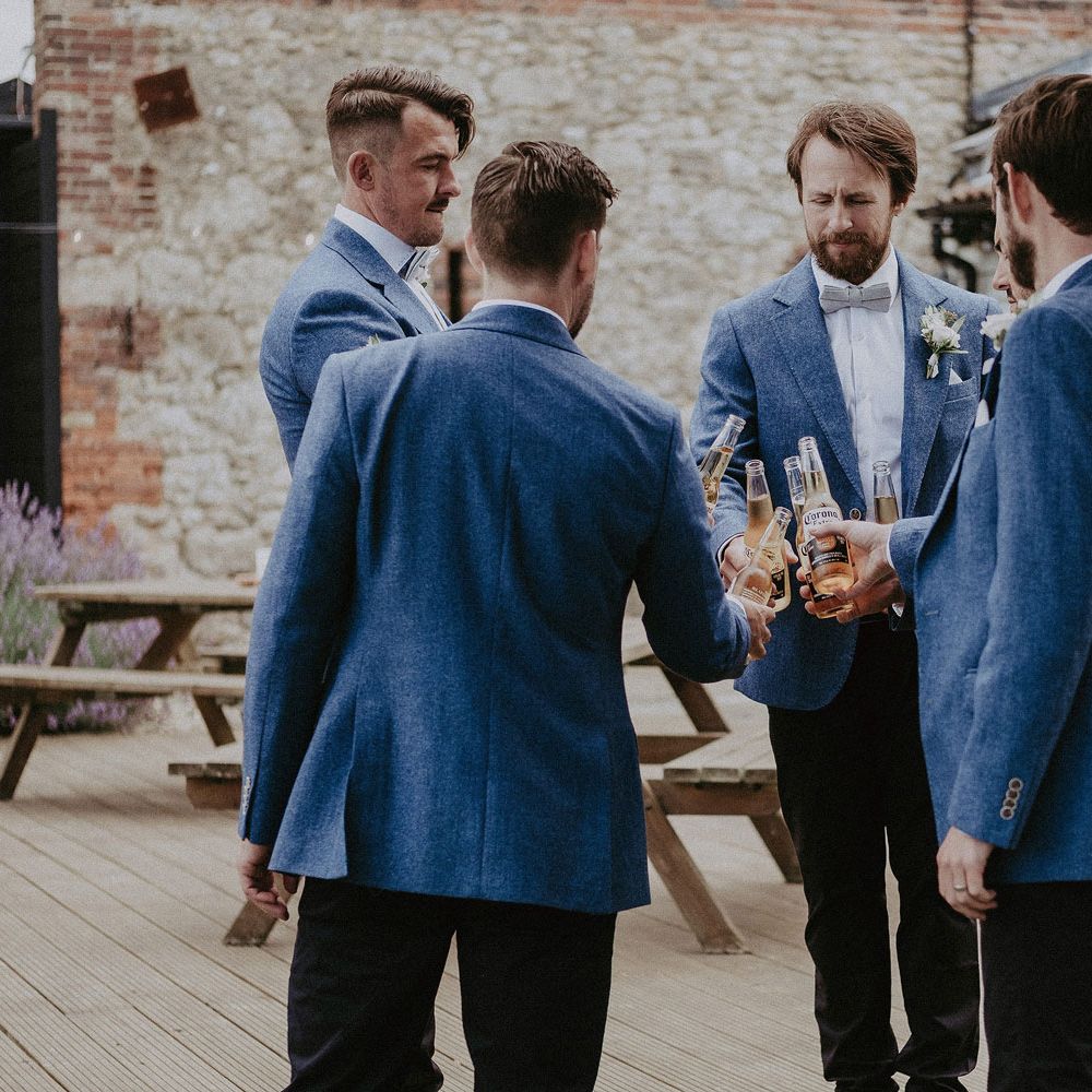 Groomsmen in blue suit jackets and bow ties all clink bottles of Corona before Isle of Wight wedding with macrame wedding decor