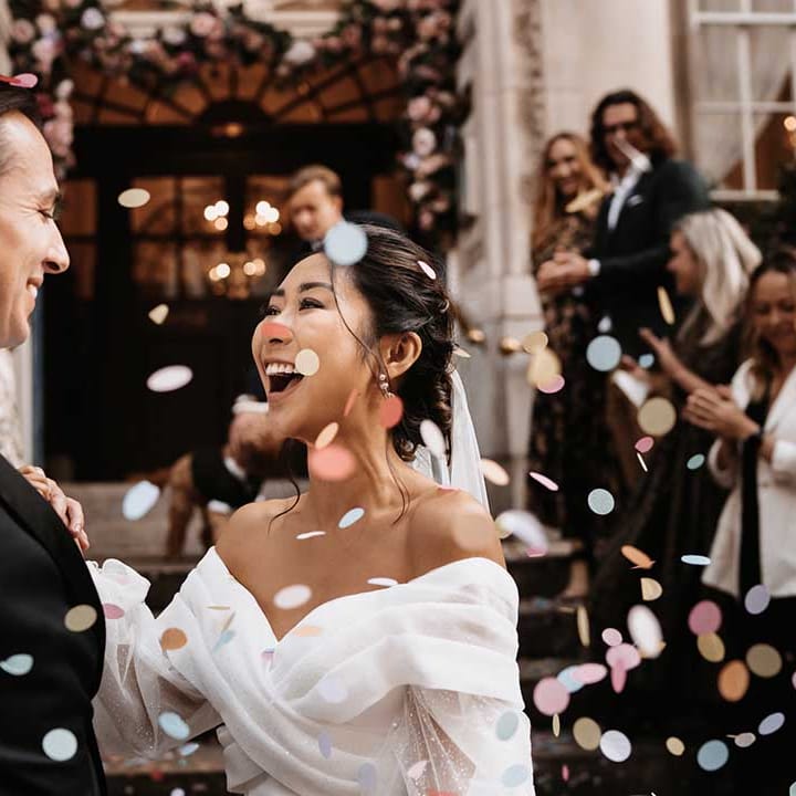 Bride and Groom smile with Joy outside town hall with pastel colour confetti as wedding guests cheer
