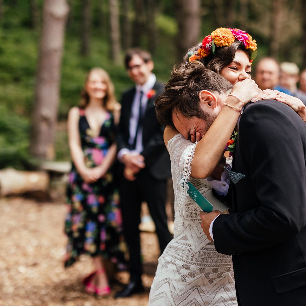 Bride in off the shoulder Grace Loves Lace wedding dress and flower crown hugs groom and he holds vows during woodland wedding ceremony