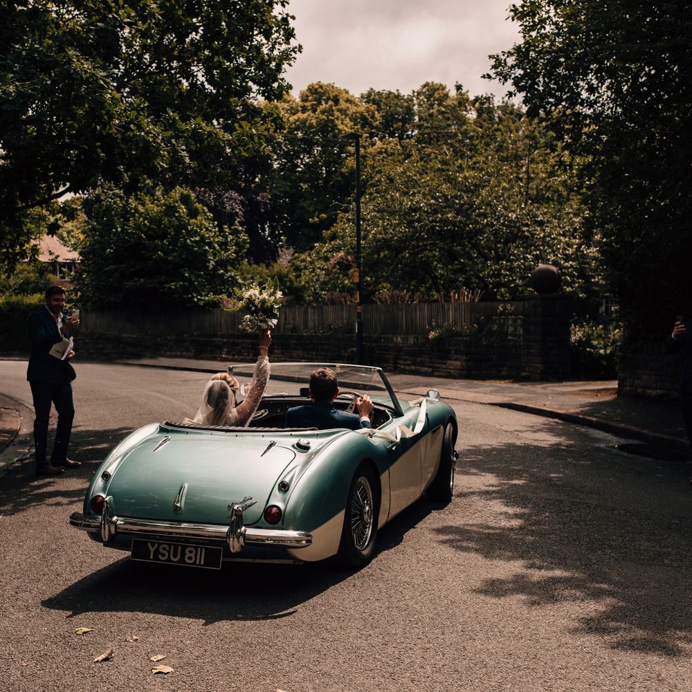 Bride holds up bouquet in the air and groom drives them both away from the church in vintage blue and white sports car after wedding ceremony