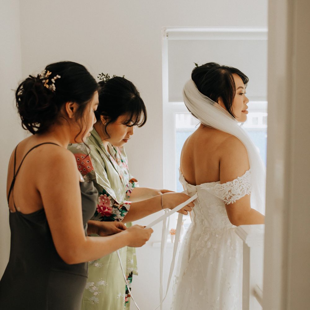 Bride in bardot corset back wedding dress and veil has the back of her dress tied by two bridesmaids before wedding