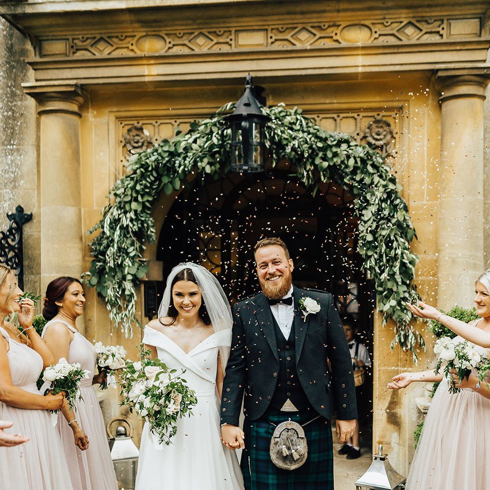 Confetti moment with Bride in a Suzanne Neville princess wedding dress walking out of Foxhill Manor with her groom in a tartan kilt surrounded by their wedding party in pink bridesmaid dresses 