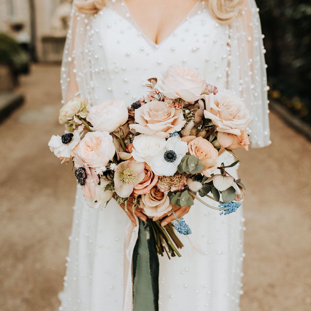 Bride in white wedding dress with pearl mesh overlay stands outside holding mixed pastel wedding bouquet with roses and anemones