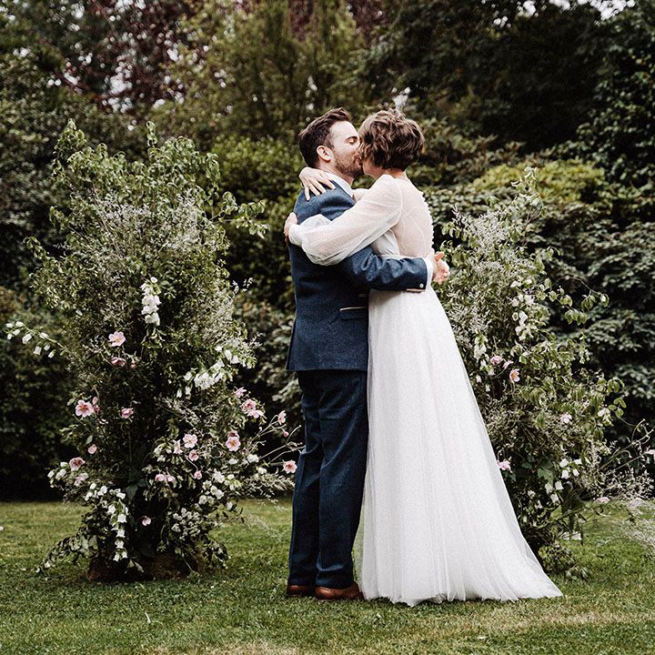 Groom in blue suit and gold patterned tie with handkerchief kisses bride in long sleeve wedding dress in front of flower columns
