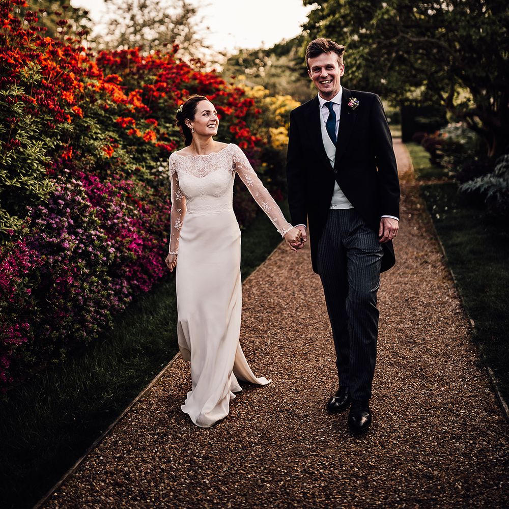 Groom in morning suit and bride in satin wedding dress walk around the gardens of Wadhurst Castle 