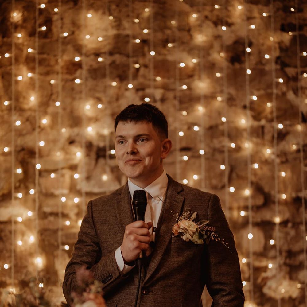 Groom reads out his speech in three piece brown suit with fairy light backdrop at Eden Barn