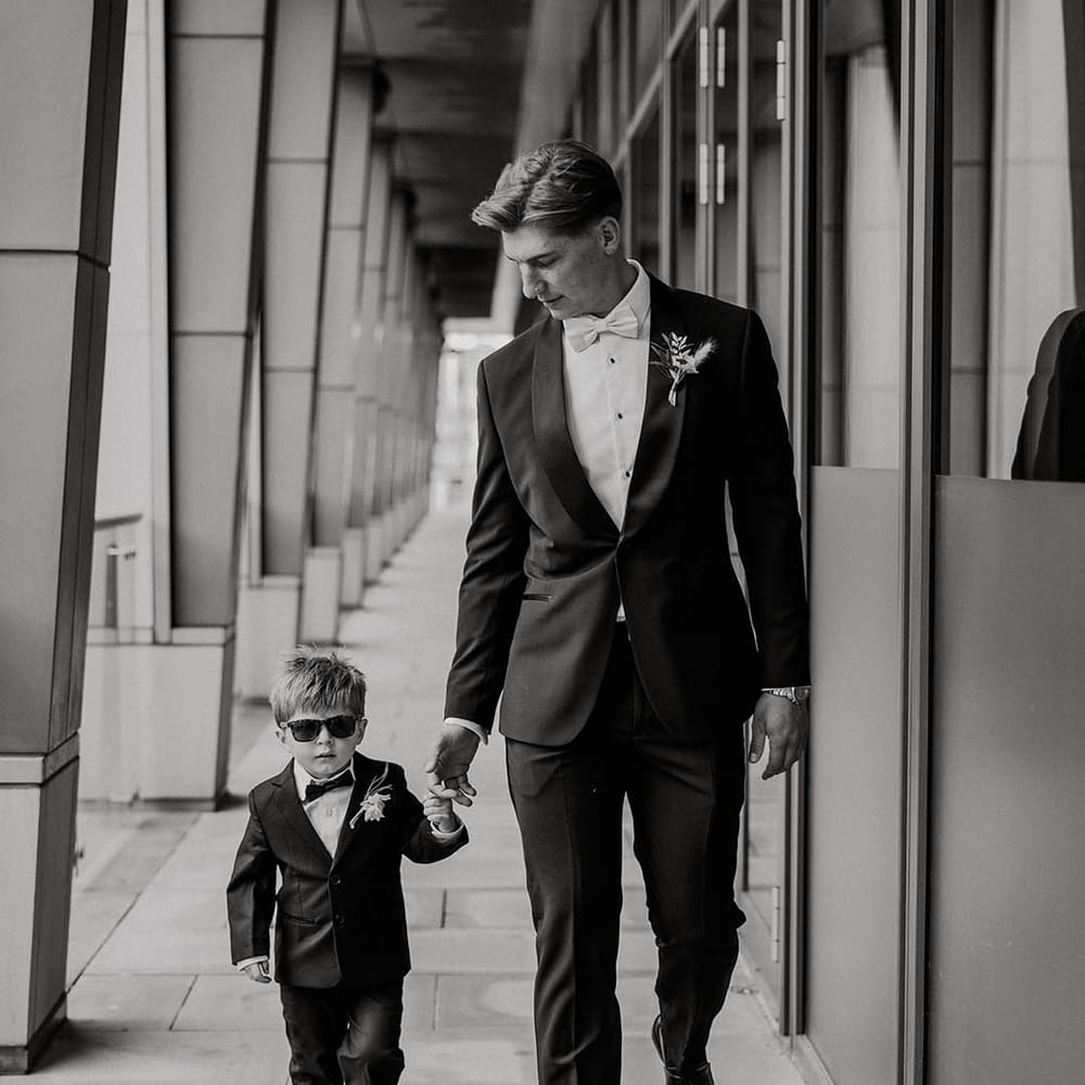 Groom in black tie with light blue bow tie walks with son in black tie and sunglasses 
