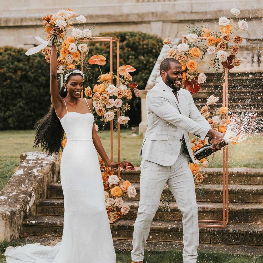 Bride in pearl strappy wedding dress and groom in grey checkered suit doing champagne pop