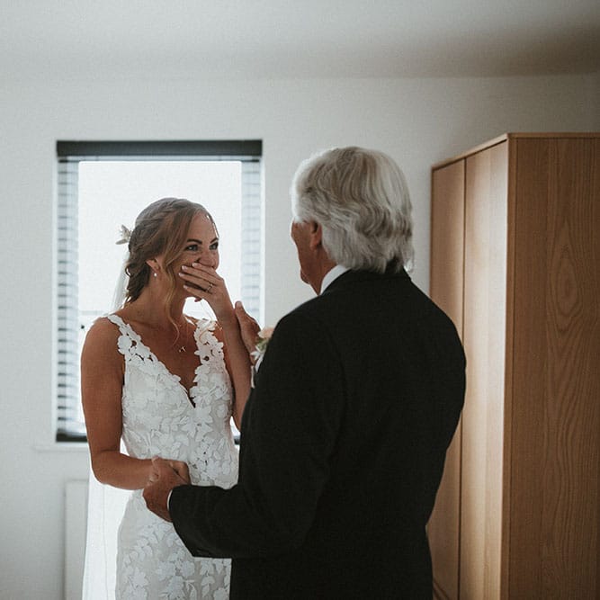 Bride wearing lace Enzoani wedding dress becomes emotional with her father on the morning of her wedding day