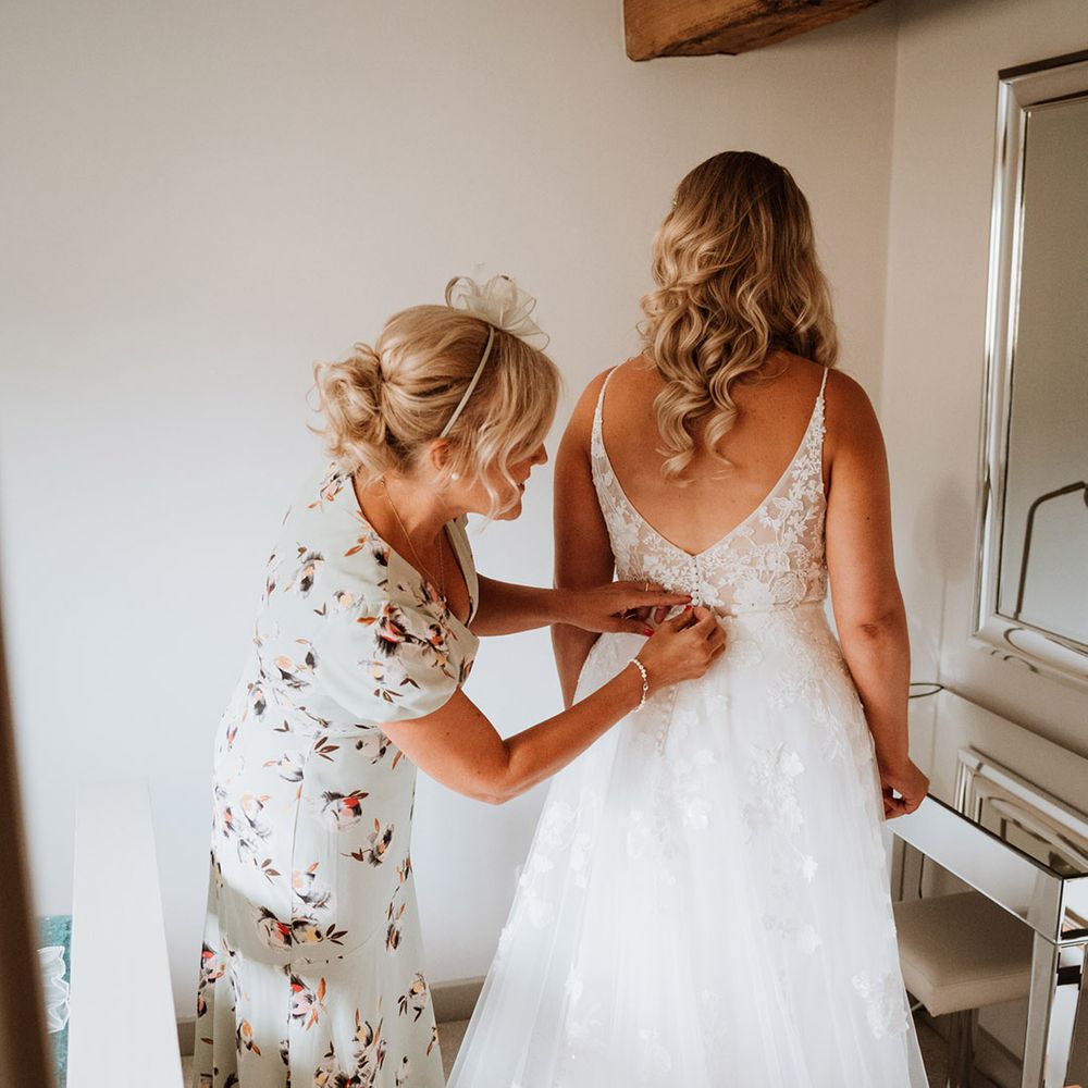 Bride in a low back floral lace wedding dress is buttoned up by the mother of the bride in a floral dress 