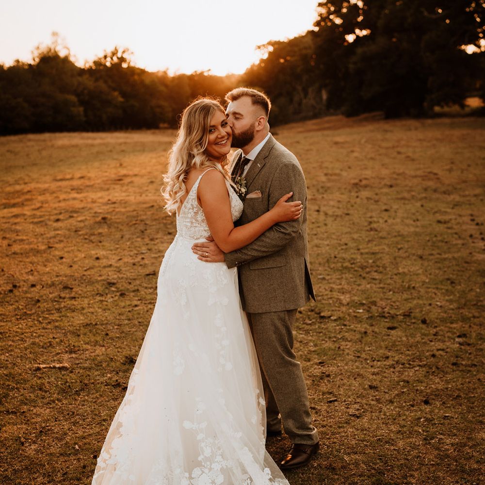 The groom in a grey tweed suit kisses the bride in a flower lace wedding dress on the cheek 