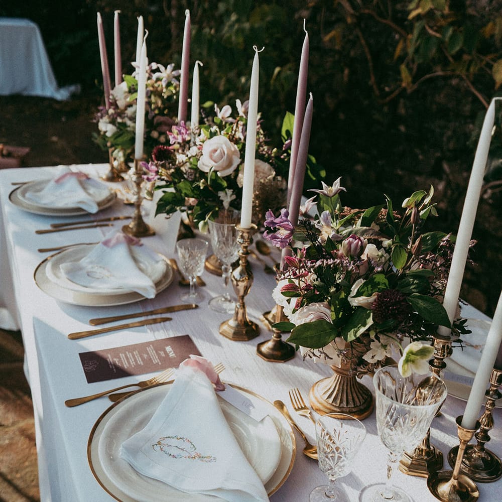 Wedding tablescape with lavender and white tapered candles in gold candle holders, mixed lavender, neutral and white bouquets, embroidered white napkins and gold cutlery 